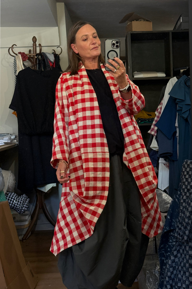 A woman wearing a red and white gingham check longline cardigan over a black top and dark gray pants, taking a mirror selfie in a cluttered clothing room.